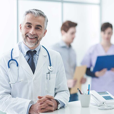 Älterer Arzt mit grauen Haaren und Stethoskop lächelt freundlich in die Kamera. Im Hintergrund sind zwei medizinische Fachkräfte, die Dokumente besprechen.